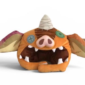 May include: A plush hat shaped like a creature with orange fabric, large ears, a pink snout, and a white horn. It features two button eyes, one blue and one green, and a wide open mouth lined with white teeth, creating a distinct character design.