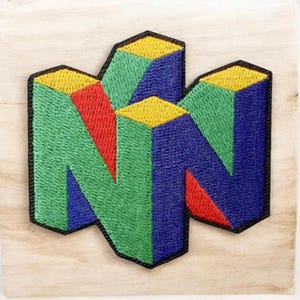 May include: A fabric patch featuring a stylized letter N in a three-dimensional block design. The embroidery uses vibrant yellow, green, blue, and red thread colors with a thick black border outlining the entire geometric shape of the letter.