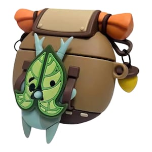 May include: A brown silicone protective case for wireless earbuds shaped like a backpack with orange straps. A light green leaf-shaped character with small horns is attached to the front. A small yellow acorn charm hangs from a black metal ring on the side.