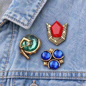 May include: Three decorative pins attached to light blue denim fabric. One pin features a red pentagonal stone in a gold setting, another has a round green stone with gold accents, and the third displays three round blue stones in a gold frame.