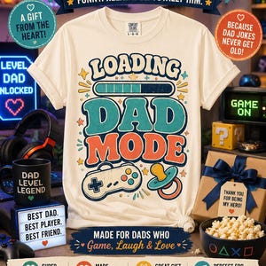 Puede incluir: Una camiseta de color crema con un estampado gráfico que incluye el texto LOADING DAD MODE, una barra de carga, un mando de videojuegos y un chupete de bebé. El diseño utiliza tipografía retro y colores como azul, naranja y verde azulado.