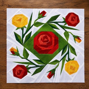 May include: A square fabric quilt block featuring a central red rose surrounded by smaller red and yellow roses, green leaves, and stems. The design is set against a light gray background with white geometric strips creating a fractured pattern.