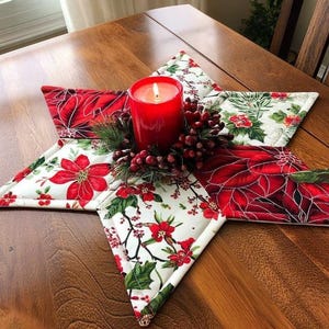 May include: A star-shaped quilted fabric mat featuring a white background with red poinsettia flowers and green holly leaves. A red pillar candle sits in the center, surrounded by a decorative ring of artificial pine needles and red berries.