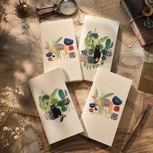 May include: Four rectangular off-white paper notebooks featuring two distinct illustrations. Two notebooks display a tan camel with a patterned saddle among red and blue flowers, while the other two feature a black cat sitting amidst green potted plants.
