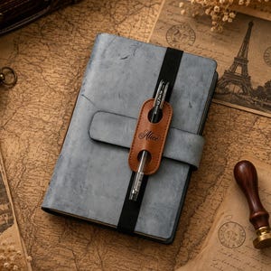 May include: A gray notebook with a distressed texture featuring a black elastic band and a tan leather pen holder strap. The strap is embossed with the name Alice and holds a clear and black gel pen. The notebook has a rectangular shape.