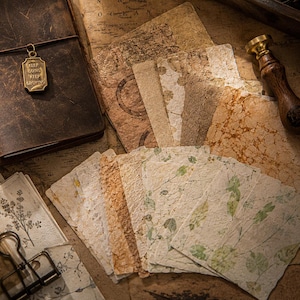 May include: A collection of textured, handmade paper sheets in various shades of beige, brown, and cream, featuring botanical prints and marbled patterns. A dark brown leather journal with a brass charm reading KEEP GOING KEEP GROWING is included.