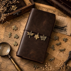 May include: A dark brown leather journal secured with an elastic band, featuring three hanging antique brass charms shaped like a dragonfly, a clover, and a butterfly. Several loose matching metal charms are scattered around the notebook.