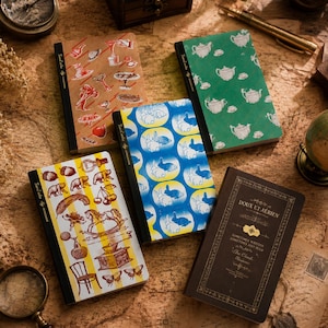 May include: Five rectangular notebooks with distinct covers: one tan with red kitchen motifs, one green with white teapots, one blue with white rabbit silhouettes, one yellow-striped with various vintage illustrations, and one dark brown with gold text.