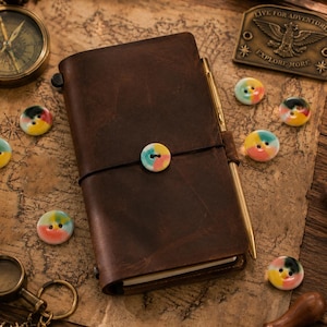 May include: A dark brown leather journal secured with a black elastic band and a multi-colored button, featuring a gold-toned metal pen tucked into the side. The journal is surrounded by several small, round, multi-colored buttons on a map background.