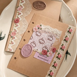 Pode incluir: Dois encartes de papel bege para agenda decorados com renda creme, pequenas rosas de croch&ecirc; cor-de-rosa e selos de cera. Um selo oval roxo tem o texto Go with the flow, e um marrom diz Dream. Uma etiqueta roxa diz Bubble Wonderland.