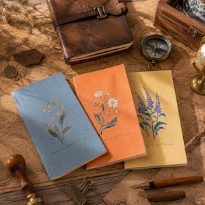 Puede incluir: Tres cuadernos rectangulares con cubiertas texturizadas en azul, naranja y amarillo, cada uno con una ilustración botánica de flores y un pequeño texto impreso. El cuaderno azul muestra lirios de los valles, el naranja margaritas y el amarillo lavanda.