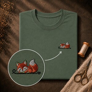 Könnte beinhalten: Ein olivgrünes T-Shirt mit einer kleinen, detaillierten Stickerei eines schlafenden Fuchses auf der Brust. Der Fuchs ist orange mit einer weiß gefärbten Schwanzspitze und ruht auf einer dünnen, dunklen Linie. Ein kreisförmiger Ausschnitt zeigt den Fuchs.