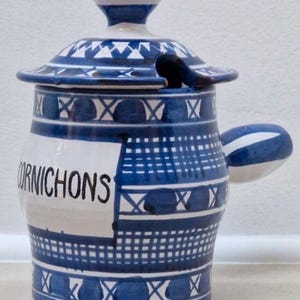 Peut inclure: Un pot en céramique avec couvercle et poignée latérale, présentant une base blanche avec des motifs géométriques peints à la main en bleu et le mot CORNICHONS imprimé en noir sur le devant. Le couvercle comporte une encoche pour ustensile.