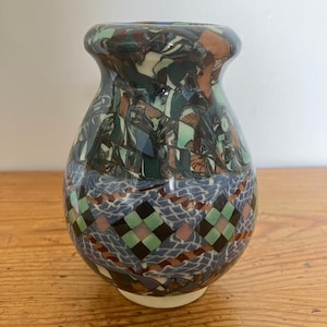 May include: A ceramic vase with a rounded body and a flared rim. The upper section features a marbled pattern in shades of dark green, blue, and brown. The lower section displays a geometric grid pattern of diamonds in green, pink, and black.