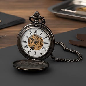 May include: A dark metallic pocket watch with a white dial featuring black Roman numerals and a visible gold-toned mechanical movement in the center. The watch has a matching dark metal chain attached to the top crown and a hinged cover.