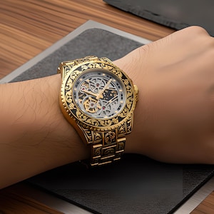 May include: A gold-tone wristwatch featuring an intricate black filigree pattern engraved on the bezel and link bracelet. The watch face displays a skeletonized dial, revealing the internal mechanical movement with visible gears and gold hands.
