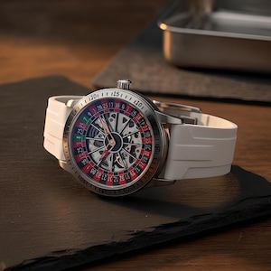 Könnte beinhalten: Eine Armbanduhr mit weißem Silikonarmband und silberfarbenem Metallgehäuse. Das Zifferblatt zeigt ein detailliertes Roulette-Rad-Design mit roten, schwarzen und grünen Segmenten, nummerierten Indizes und skelettierten Zeigern sowie einer Krone.