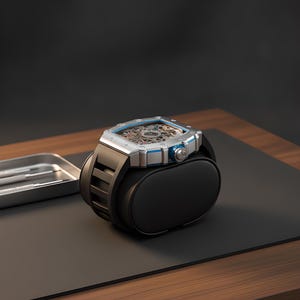 May include: A silver-toned skeleton watch with a square case and blue accents, featuring a black rubber strap. The watch is mounted on a black cylindrical display cushion, with a small metal tray containing two silver tools visible nearby.