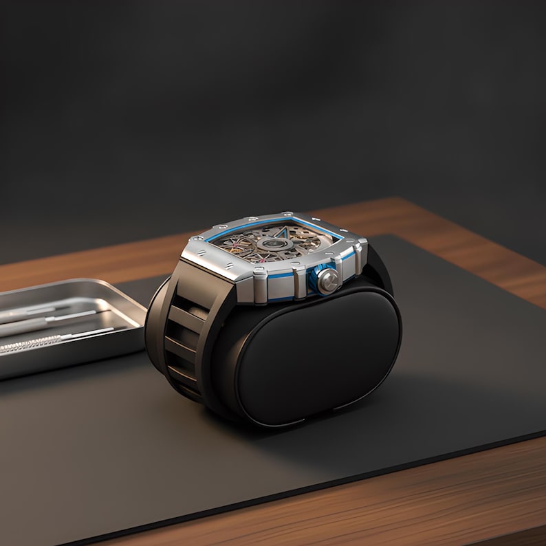 May include: A silver-toned skeleton watch with a square case and blue accents, featuring a black rubber strap. The watch is mounted on a black cylindrical display cushion, with a small metal tray containing two silver tools visible nearby.