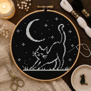 May include: A circular wooden embroidery hoop containing a black fabric piece with a white cross-stitch design of a stretching cat under a crescent moon and stars. A silver needle with white thread is attached to the fabric near the cat's tail.