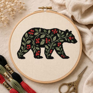 May include: A circular wooden embroidery hoop containing a cross-stitch pattern of a black bear silhouette filled with green leaves and small red berries on a cream-colored fabric background, featuring a metal tension screw at the top.