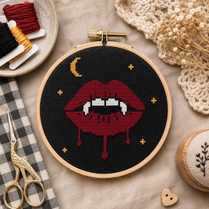 May include: A circular wooden embroidery hoop containing a cross-stitch design of red lips with white fangs on a black background. The design includes a yellow crescent moon and small yellow stars. The hoop is surrounded by embroidery supplies.
