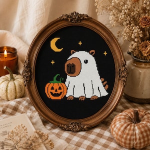 May include: A cross-stitch embroidery in an oval wooden frame featuring a brown capybara wearing a white ghost costume. The design includes an orange jack-o'-lantern, a yellow crescent moon, and small yellow stars against a black background.