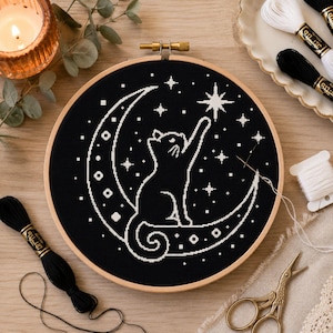 May include: A circular wooden embroidery hoop containing a black fabric piece with a white cross-stitch design of a cat sitting on a crescent moon and reaching for a star. The design includes various small stars and geometric patterns.