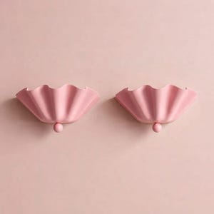 Peut inclure: Deux appliques murales identiques rose clair présentant un design large et ondulé en forme de coquillage. Chaque luminaire possède une finition mate lisse et un petit détail en forme de boule sphérique à la base de l'abat-jour plissé.