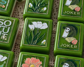 May include: A collection of rectangular green tiles featuring raised relief designs. The tiles display white floral illustrations, pink flowers, and a stylized figure labeled JOKER, alongside tiles with the text SOUTH and directional arrows.