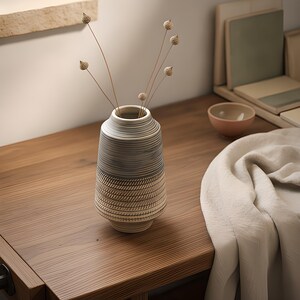 May include: A cylindrical ceramic vase with a tapered neck, featuring a two-tone design with a smooth grey upper section and a textured lower section with diagonal etched lines, containing five thin, dried stems with small round seed pods.