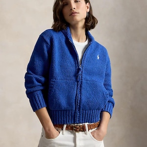May include: A bright blue zip-up knit jacket with a high collar, long sleeves, and ribbed cuffs and hem. The jacket features a small white embroidered logo on the left chest, a full-length front zipper, and two front pockets. It is worn over a white shirt.