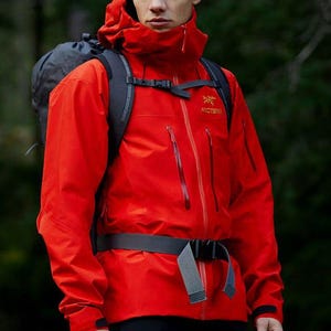 以下が含まれることがあります： ハイカラー、調節可能な袖口を備えた長袖、胸元の垂直ジッパー付きポケットが特徴の鮮やかな赤色のテクニックフード付きジャケット。左胸にはArc'teryxのロゴが刺繍されています。ダークグレーのバックパックを背負っています。