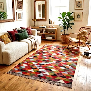 Puede incluir: Una alfombra rectangular con un patrón geométrico repetitivo de triángulos en tonos rojo, naranja, verde, negro y blanco. La alfombra tiene una superficie texturizada y detalles de flecos cortos a lo largo de los dos bordes más cortos.