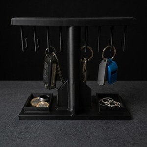May include: A black T-shaped organizer stand featuring a horizontal bar with multiple hooks for hanging keys and key fobs. The base includes two integrated rectangular trays for storing small items like coins, rings, and other accessories.