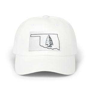 May include: A white baseball cap featuring a rectangular patch on the front. The patch displays a black outline of the state of Oklahoma with a detailed, textured black and white illustration of an arrowhead centred within the state shape.