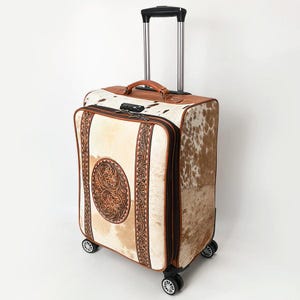 May include: A rolling suitcase featuring a brown and white cowhide pattern with tan leather trim. The front panel displays a circular embossed floral design between two vertical tooled leather strips. It includes a retractable handle and wheels.
