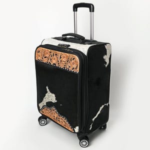 May include: A rolling suitcase featuring a black and white cowhide pattern, accented with tan leather panels displaying intricate floral tooling. The luggage includes a retractable handle, a top carry handle, a combination lock, and four wheels.