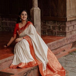 May include: A cream-coloured saree featuring a repeating geometric pattern and a wide, vibrant orange border with intricate gold metallic floral embroidery. The matching blouse is solid red with gold-toned sleeve cuffs and a gold-toned necklace.