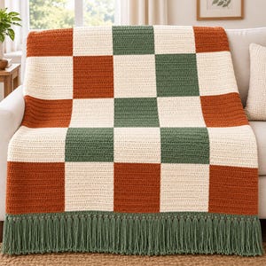 Pode incluir: Uma manta de crochê com um padrão xadrez de quadrados alternados em laranja queimado, verde floresta e creme. A manta é finalizada com uma franja longa e densa em verde floresta ao longo da borda inferior do tecido.