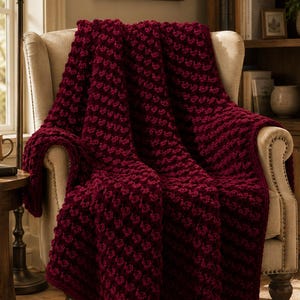 Op de afbeelding: Een diepbordeauxrode, grof gebreide plaid met een gestructureerd, herhalend noppenpatroon. De deken is gedrapeerd over een beige gestoffeerde oorfauteuil, waarbij de dikke garenconstructie en de rijke, donkerrode kleur goed zichtbaar zijn.