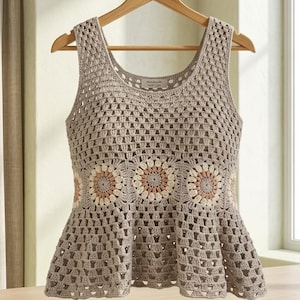 May include: A sleeveless crochet top in a light taupe color featuring a scoop neckline and a peplum hem. The garment is constructed with a granny square stitch pattern and includes a horizontal band of circular floral motifs in cream and dusty rose.