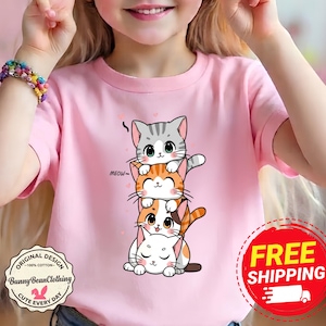 May include: A light pink t-shirt featuring a graphic of four stacked cartoon kittens in gray, orange, and white. The design includes the text MEOW and small pink hearts. The shirt has a round neckline and short sleeves, showcasing a playful illustration.