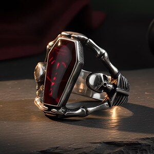Op de afbeelding: Een zilverkleurige metalen ring met een centraal doodskistvormig element met een dieprode doorschijnende inleg. De ringband is ontworpen met skeletachtige handmotieven die de kist lijken vast te houden, inclusief kleine schedeldetails aan de zijkanten.