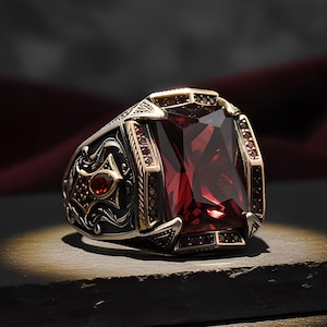 May include: A silver-toned ring featuring a large, rectangular, deep red faceted gemstone at the center. The band is decorated with intricate, dark-oxidized metal scrollwork and smaller red stone accents along the sides and the bezel setting.
