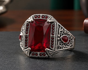 May include: A silver ring featuring a large rectangular faceted red gemstone in the center, surrounded by smaller red stones and intricate metal filigree patterns on the band, including a teardrop-shaped red stone on the side.