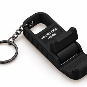 May include: A black plastic foldable phone stand keyring featuring a metal split ring. The device has a rectangular body with a textured surface and a hinged support arm. The text "YOUR LOGO HERE" is printed in white on the top section of the stand.