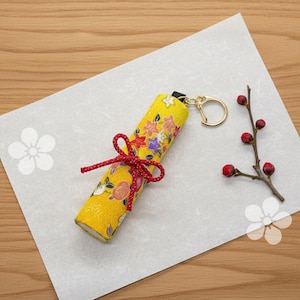 May include: A cylindrical fabric keychain featuring a bright yellow background with a colorful floral pattern, tied with a red braided cord. The keychain includes a gold-toned metal ring and is displayed alongside a small branch with red berries.