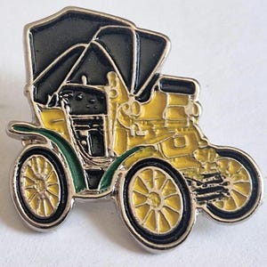 Puede incluir: Un pin de esmalte metálico con forma de automóvil antiguo con carrocería amarilla, techo negro y guardabarros verdes. El diseño presenta ruedas de radios detalladas, una parrilla delantera prominente y un estilo clásico de coche descapotable.
