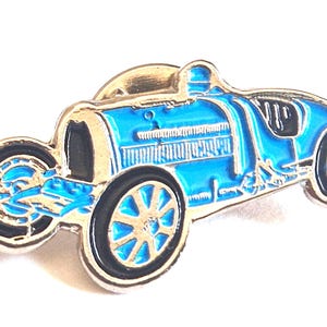Puede incluir: Un pin de esmalte metálico con forma de coche de carreras antiguo. El coche está acabado en un color azul brillante con contornos y detalles en metal plateado. Presenta un diseño clásico de cabina abierta, ruedas de radios y un capó largo con rejillas.